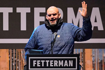 John Fetterman giving a speech wearing a blue quilted jacket and raising his left hand