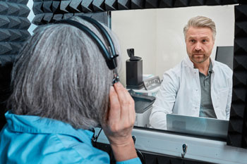 Audiologist doing hearing check-up to gray-haired woman in soundproof audiometric booth with audiometer.