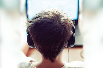 Viewed from behind, a boy wearing headphones sits in front of a computer screen.