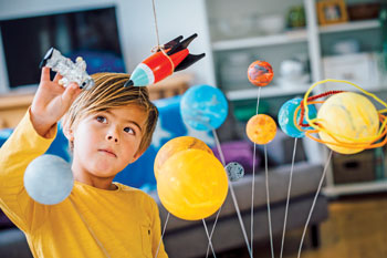 Boy in a yellow shirt plays with a space explorer and rocketship amid a model of the solar system.