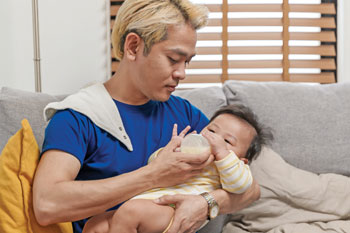 Asian man with dyed blond hair gives an infant a bottle.